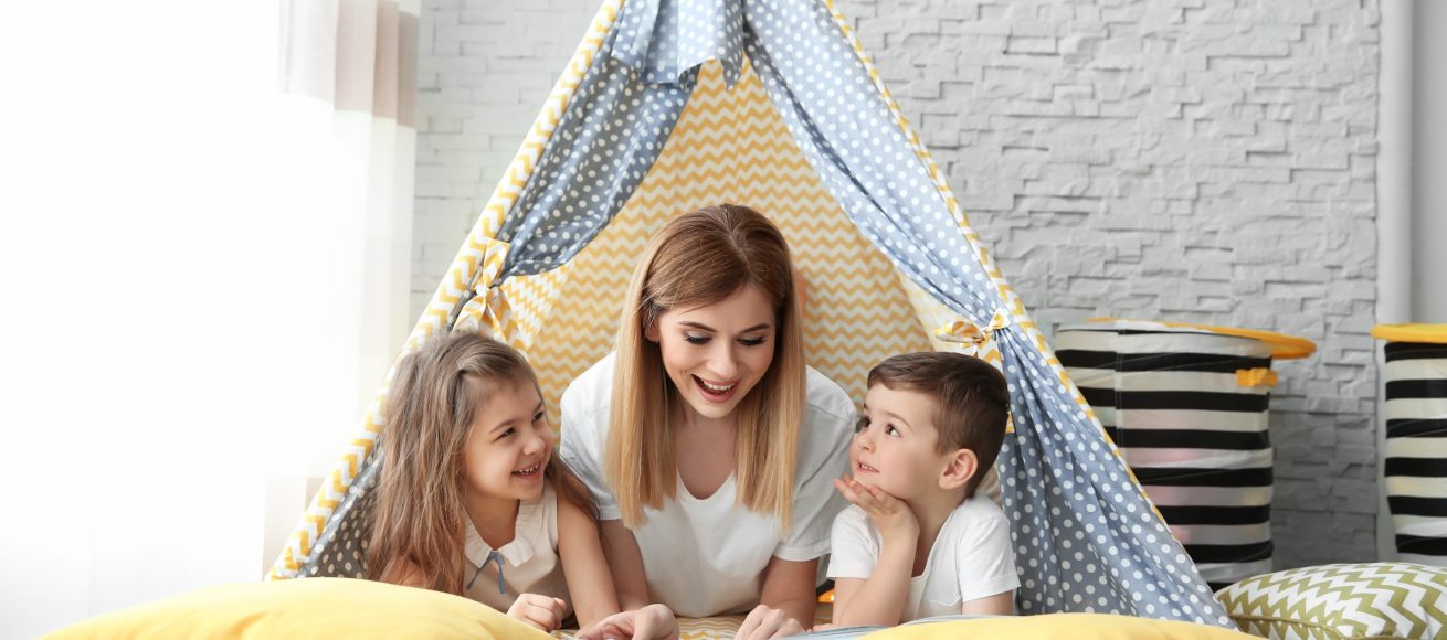 Nanny and little children reading book in tent at home